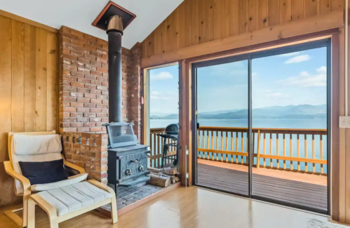 Cozy interior with a brick wall, wood stove, and large windows showcasing a scenic lake view.